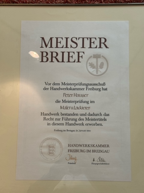 Innenansicht einer Lagerhalle mit mehreren Regalen, die mit Baumaterialien bestückt sind. Es gibt Säcke mit Beschriftungen, Eimer, Werkzeuge und diverse andere Bauartikel. Die Regale sind in verschiedenen Farben, darunter Gelb und Blau. Der Boden der Halle ist betoniert und mit gelben Markierungen versehen. Das Dach besteht aus Metallträgern, und es gibt mehrere Beleuchtungskörper, die die Halle erhellen.